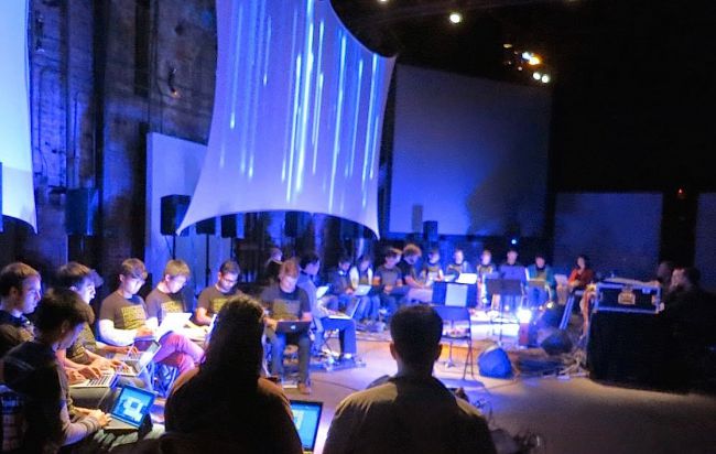 An image of several students using their laptops to form an orchestra on a grand stage.